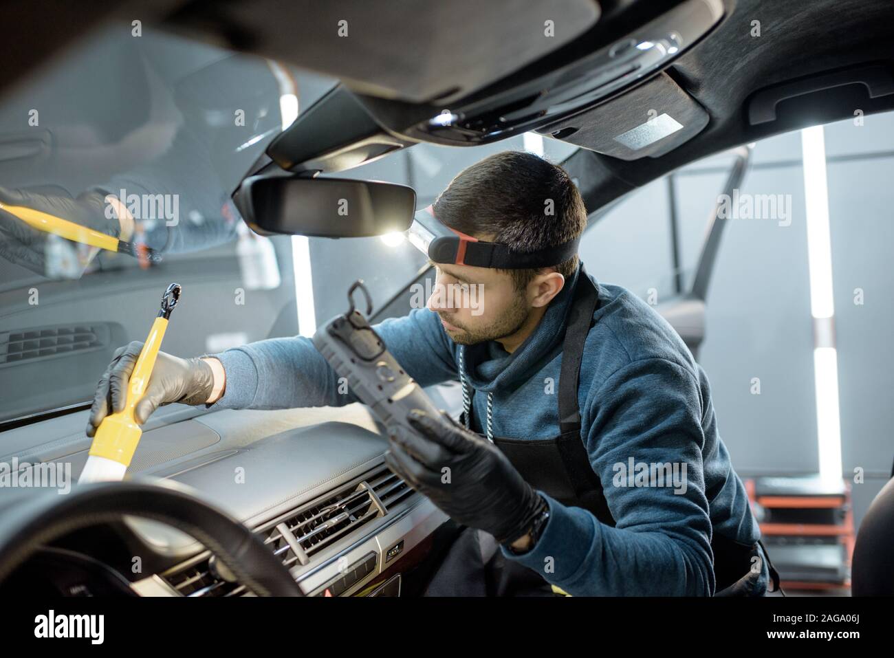 Car service worker in uniform provides a professional vehicle interior cleaning, wiping front panel with a brush at the car service station Stock Photo