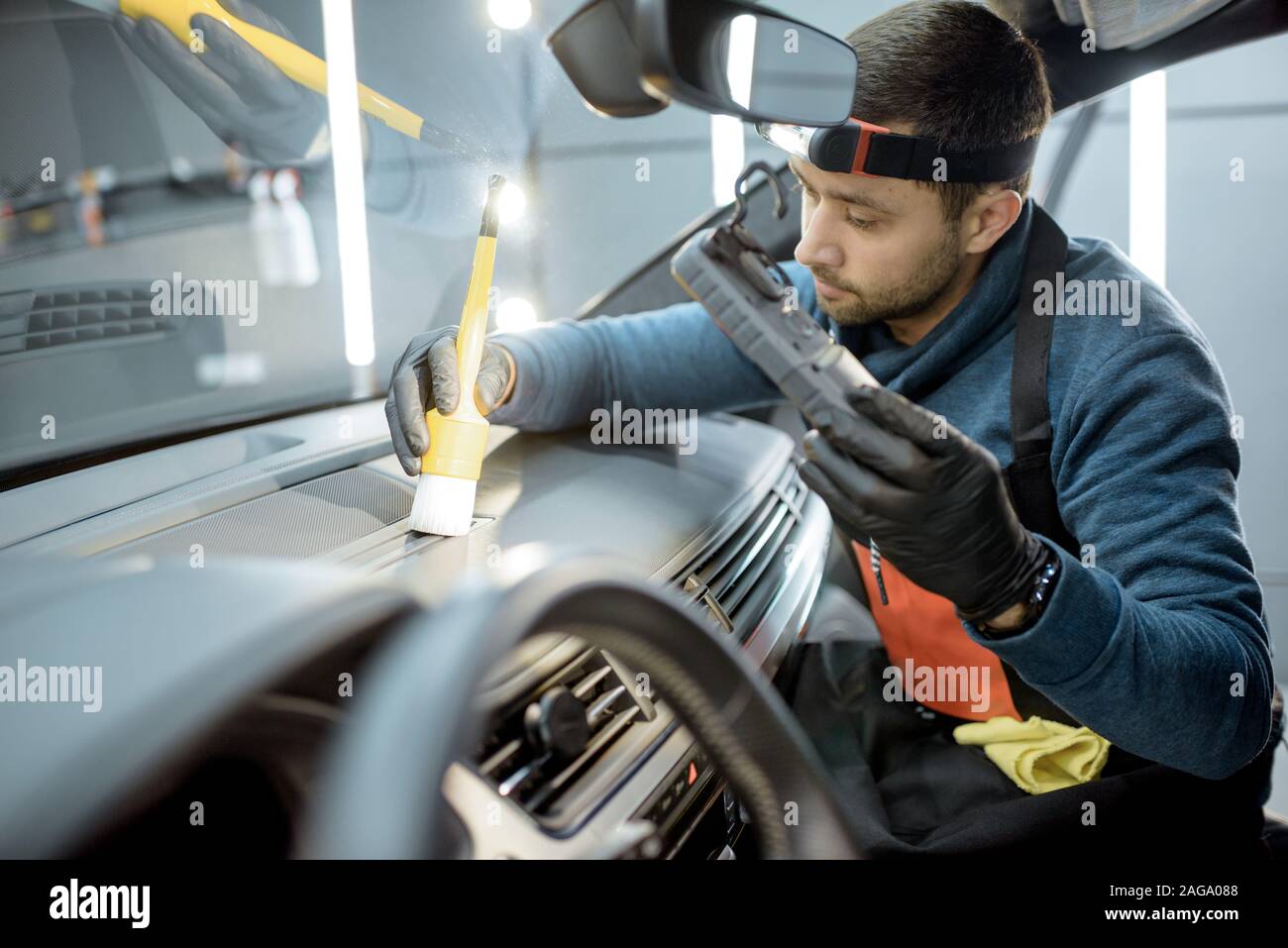 Car service worker in uniform provides a professional vehicle interior cleaning, wiping front panel with a brush at the car service station Stock Photo