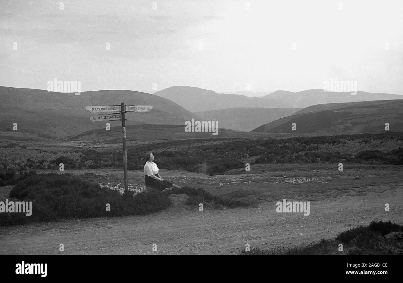 1950s, historical, a lady walker in the Shropshire hills at 'Long Mynd' sitting by a road sign for Ratlinghope, Woolstaston and Church Stretton, Shropshire, England. A heath and moorland plateau, in 1965 large areas of Long Mynd - almost all its upland area - were brought by the National Trust and designated as an area of outstanding natural beauty (ANOB). Stock Photo