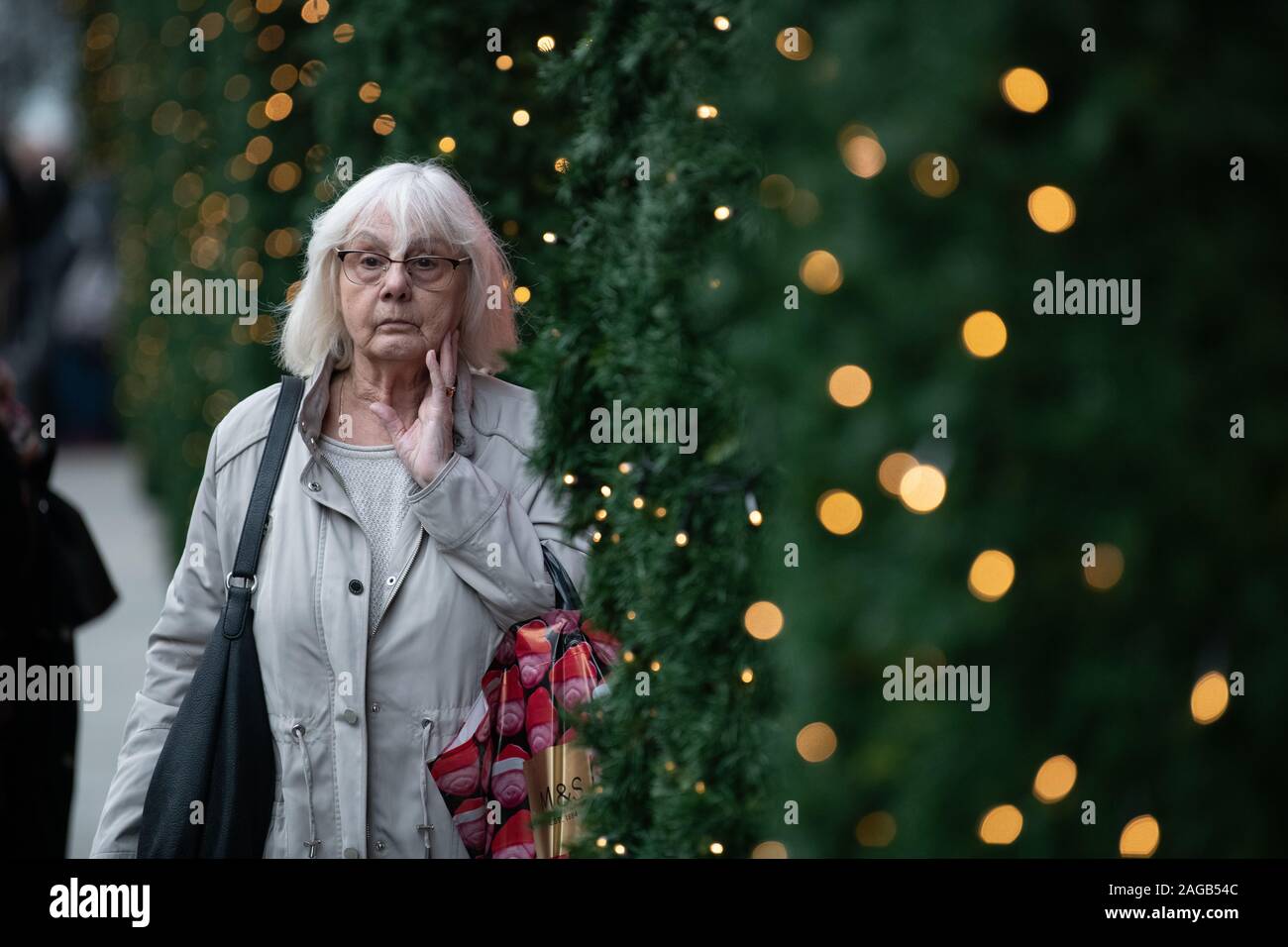 Grey hair lady walking deep in thoughts with Christmas trees and lights outside Selfridges. She is caring M&S bag Stock Photo