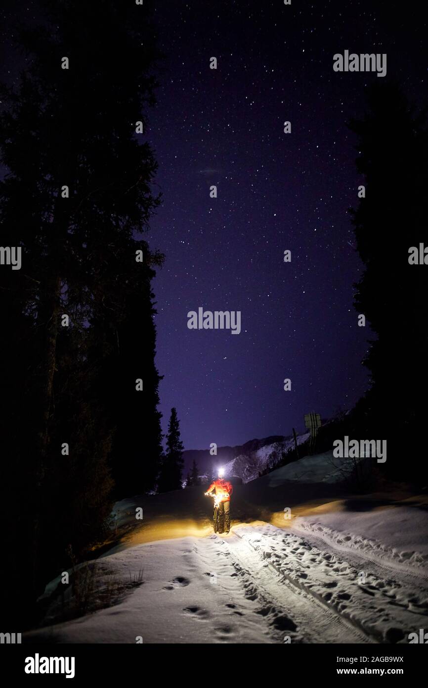 Man in red jacket with bicycle at winter snowy forest in the mountains under night sky with stars Stock Photo