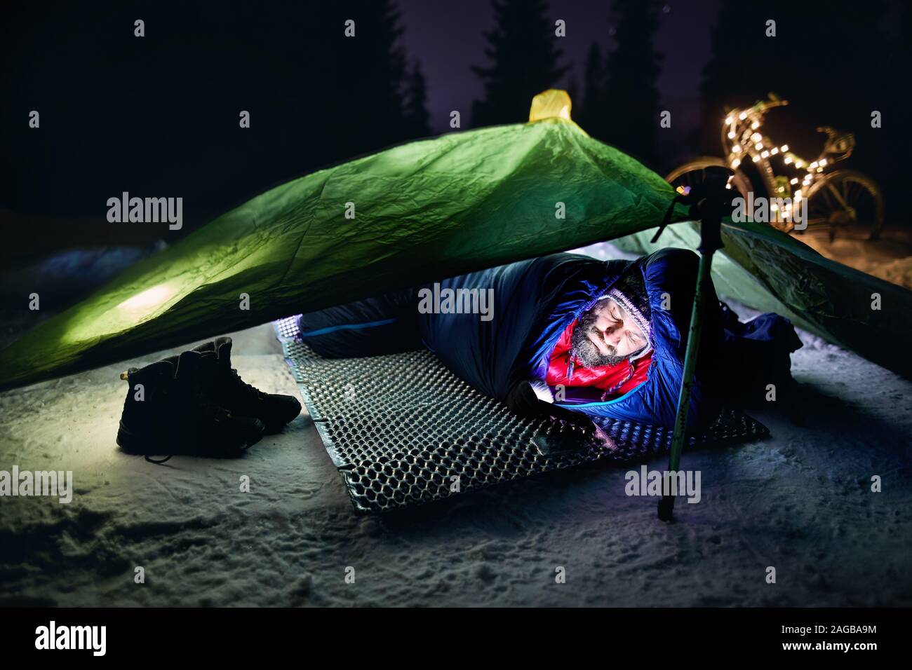 Bearded man in tent in sleeping bag is looking to his phone at winter night. Bicycle decorated with Christmas lights at background. Bikepacking and Ch Stock Photo