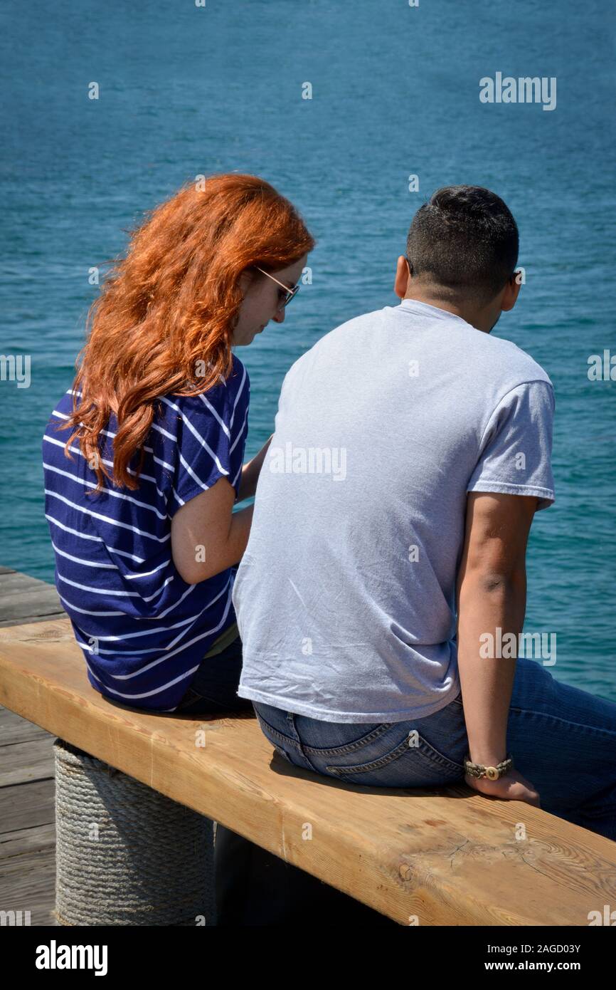 A rear view of a red headed young woman with long hair sitting alongside a young man on a wooden bench on a pier overlooking the Pacific Ocean Stock Photo