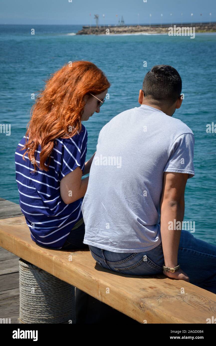 A rear view of a red headed young woman with long hair sitting alongside a young man on a wooden bench on a pier overlooking the Pacific Ocean Stock Photo