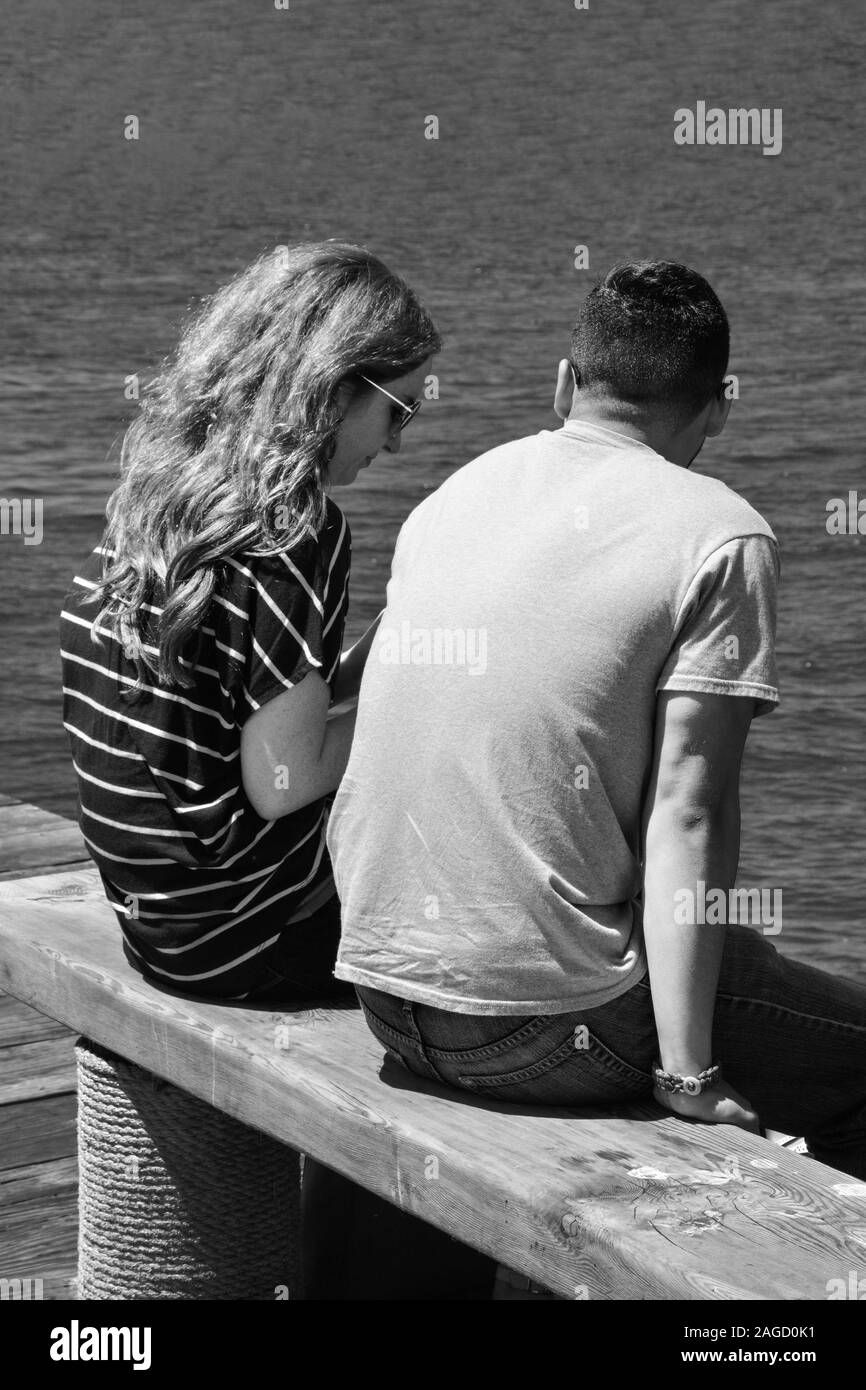 A rear view of a  young woman with long hair sitting alongside a young man on a wooden bench on a pier overlooking the Pacific Ocean in Southern CA Stock Photo