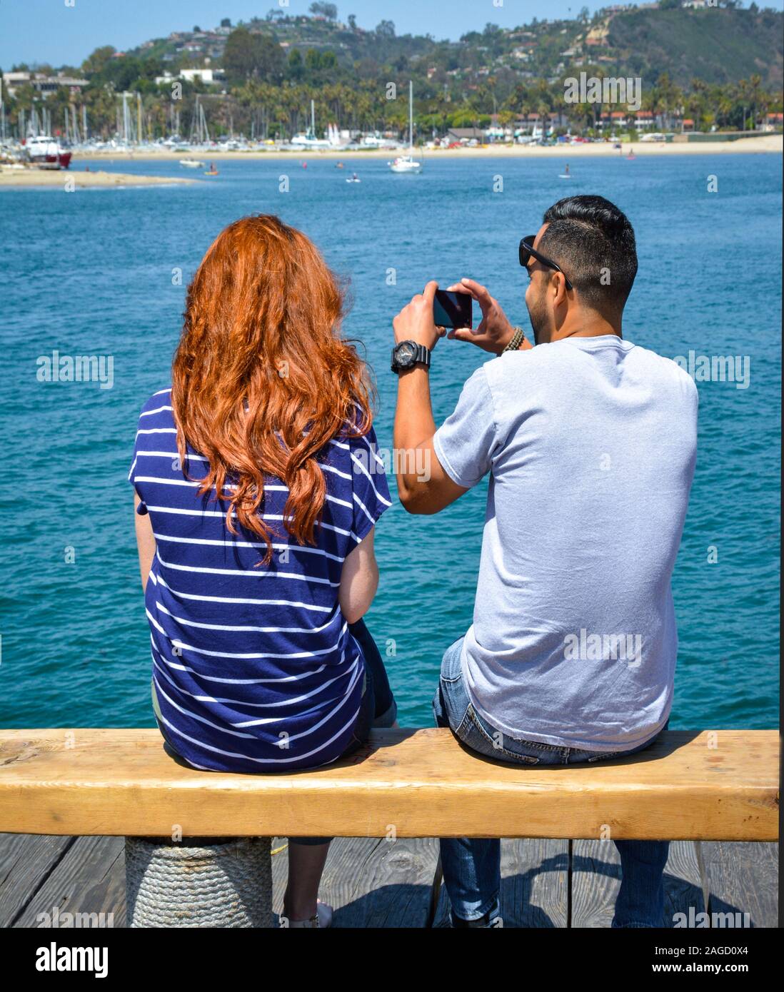 A rear view of a seated red headed young woman sitting alongside a young man using his camera phone from a pier at the harbor in Santa Barbara, CA Stock Photo
