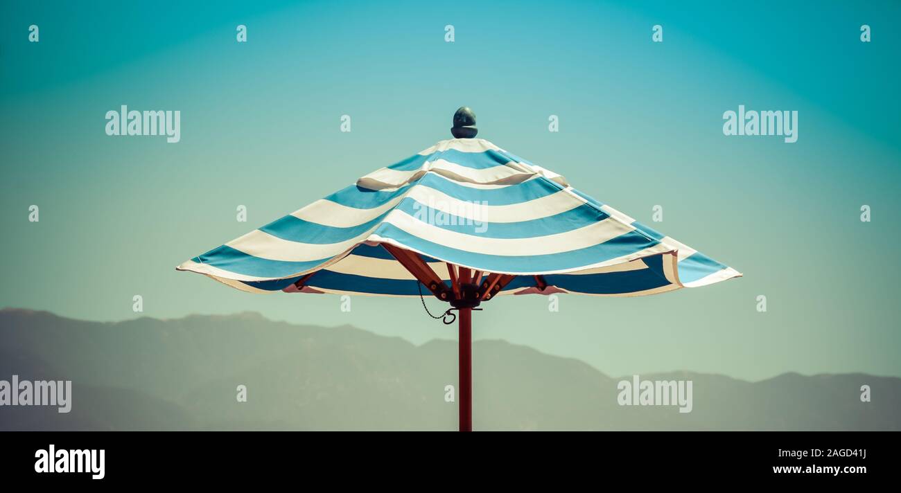 A cropped panoramic format view of sky and a lone blue striped sun umbrella with atmospheric foggy mountain range background in Southern California Stock Photo