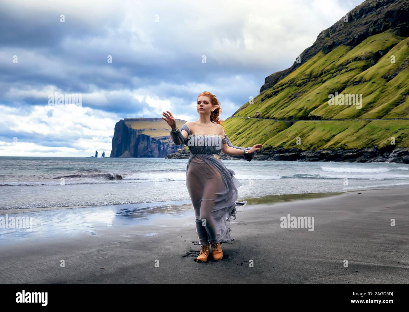 A young  woman dressed as an elf stays on the beach. Two sea stacks (Risin and Kellingin) behind. Eysturoy, Faroe Island. I have a model release Stock Photo