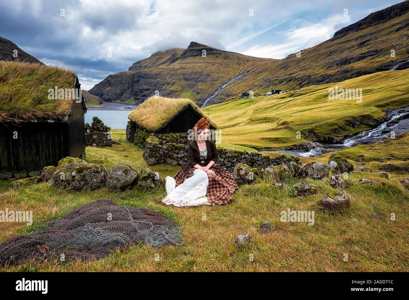 A young redhead woman sits near a stone fence in old-fashioned clothes. Saksun village. Denmark, Faroe Island. I have a model release Stock Photo