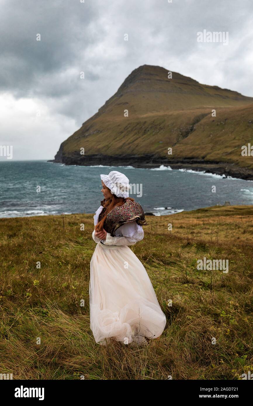 A young redhead woman stays on the grass field near Atlantic ocean coastline in old-fashioned clothes. Denmark, Faroe Island. I have a model release Stock Photo