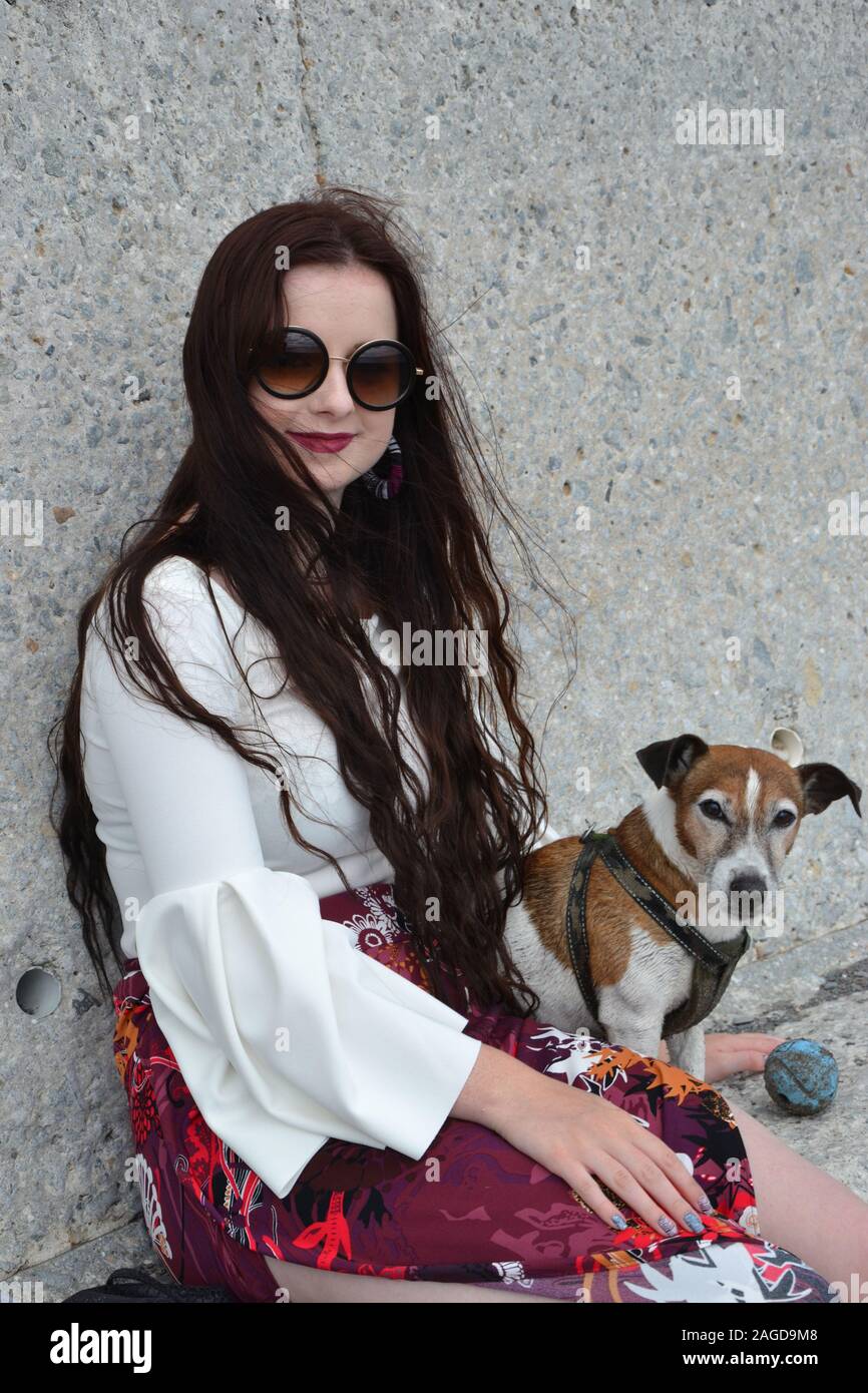 A fashionably dressed caucasian lady with long brunette hair and round sunglasses (shades) sits by a concrete wall with her white and tan Jack Russell Stock Photo