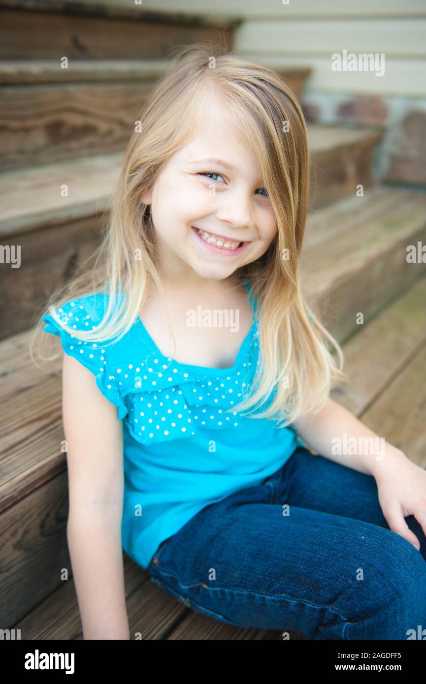 Vertical shot of a cute blonde little girl smiling naturally while sitting on the wooden stairs Stock Photo