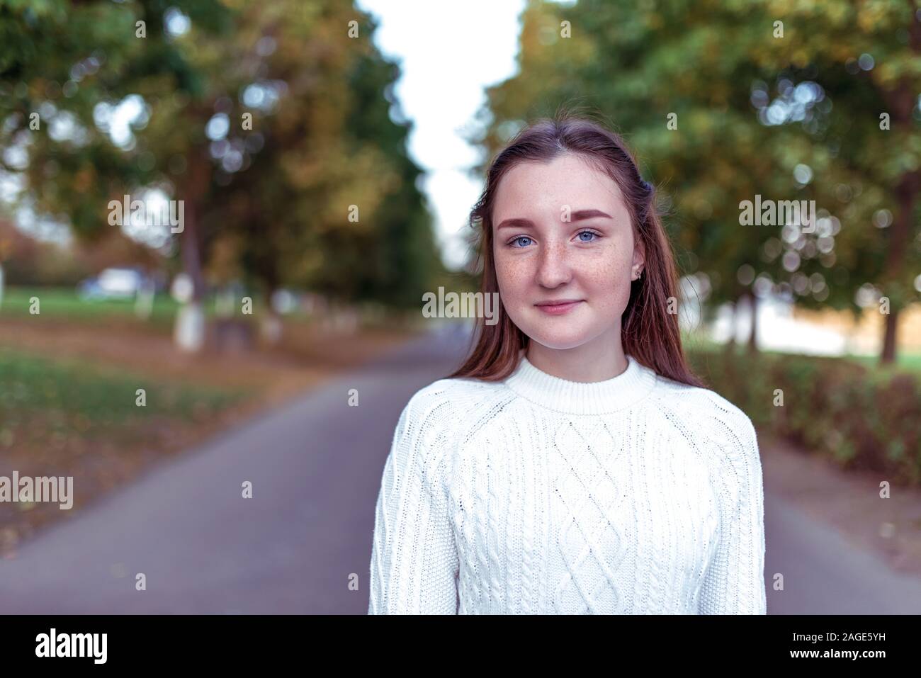 happy teenager girl of 12-15 years old, in summer in city, stands in autumn park, smiles and rejoices, in a warm white sweater. Free space for copy Stock Photo
