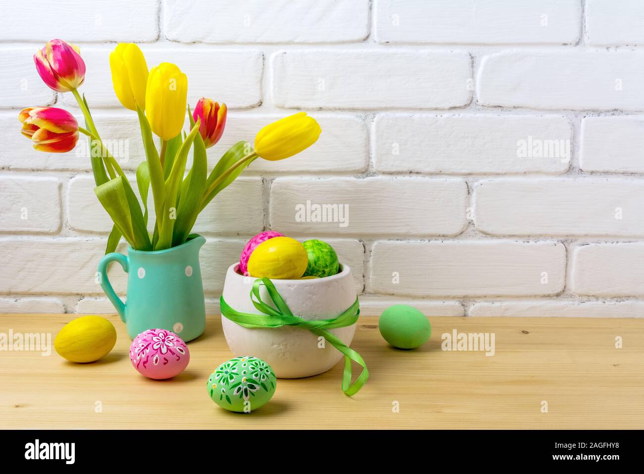 Easter rustic centerpiece with decorated eggs, green ribbon, red and yellow tulips in the polka dot mint green jug near painted brick wall Stock Photo