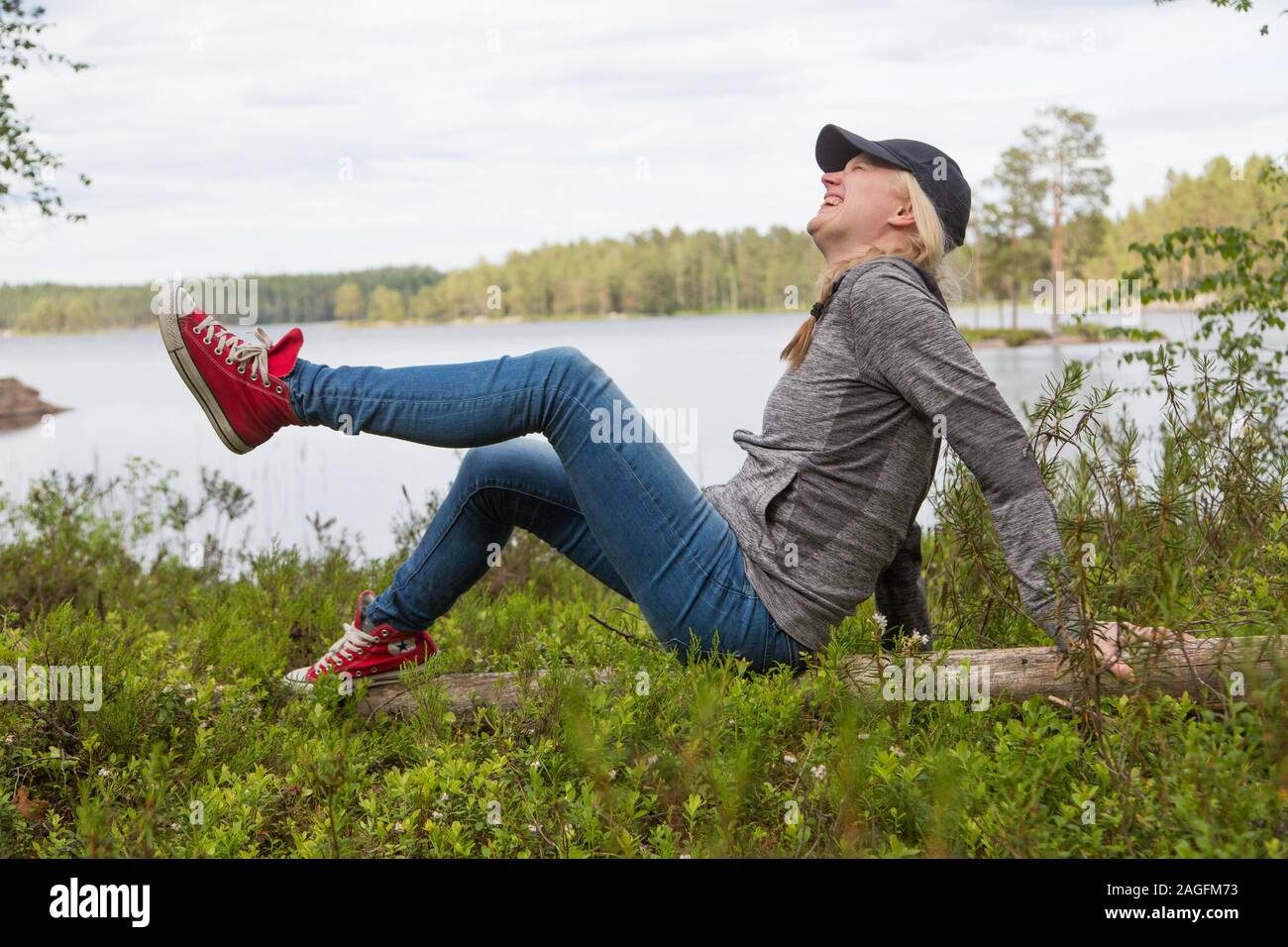 Woman in casual clothing sitting on a log by a lake dressed in converse trainers laughing Stock Photo