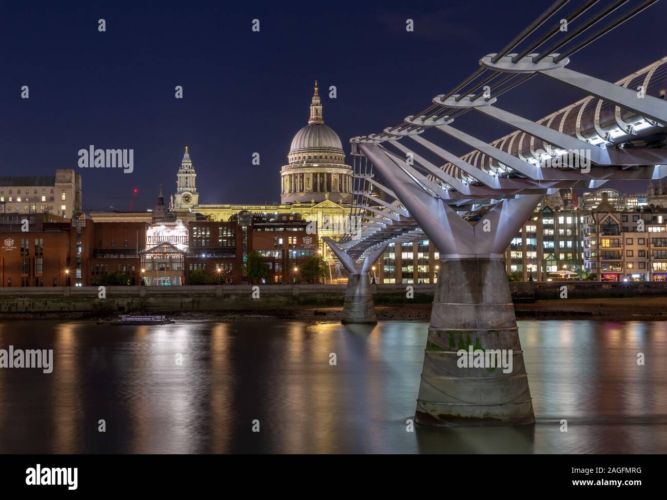 Millennium bridge surrounded by modern buildings with city lights during the night in London Stock Photo