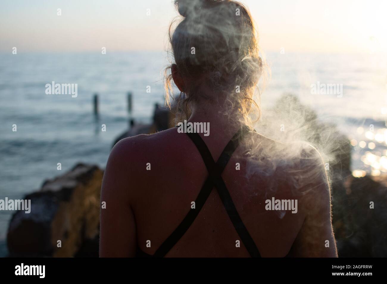 Female smoking a joint shot from behind with a blurred sea in the background Stock Photo