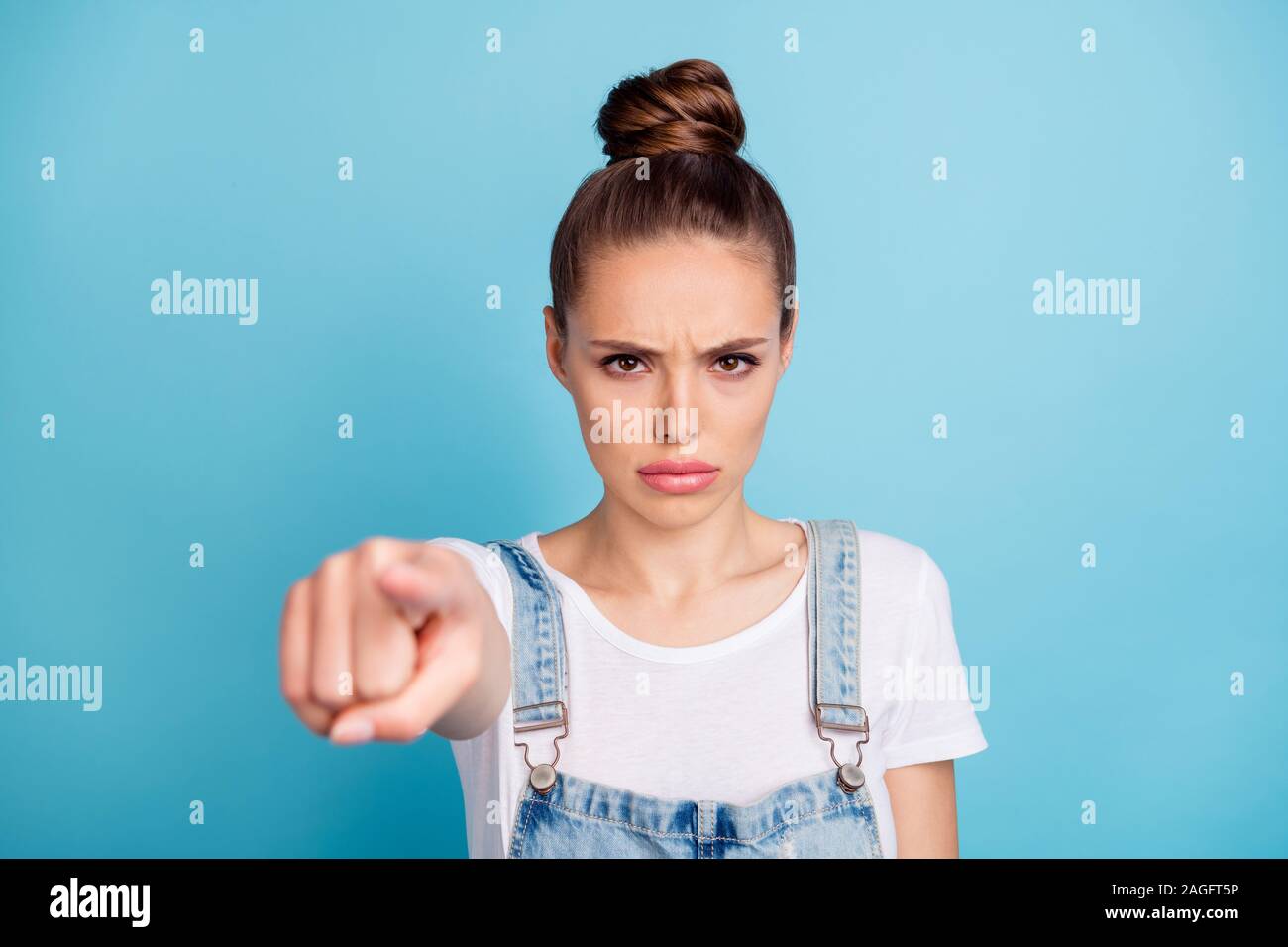 Portrait of sullen girl pointing with her index finger wearing white t-shirt denim jeans overalls isolated over blue background Stock Photo