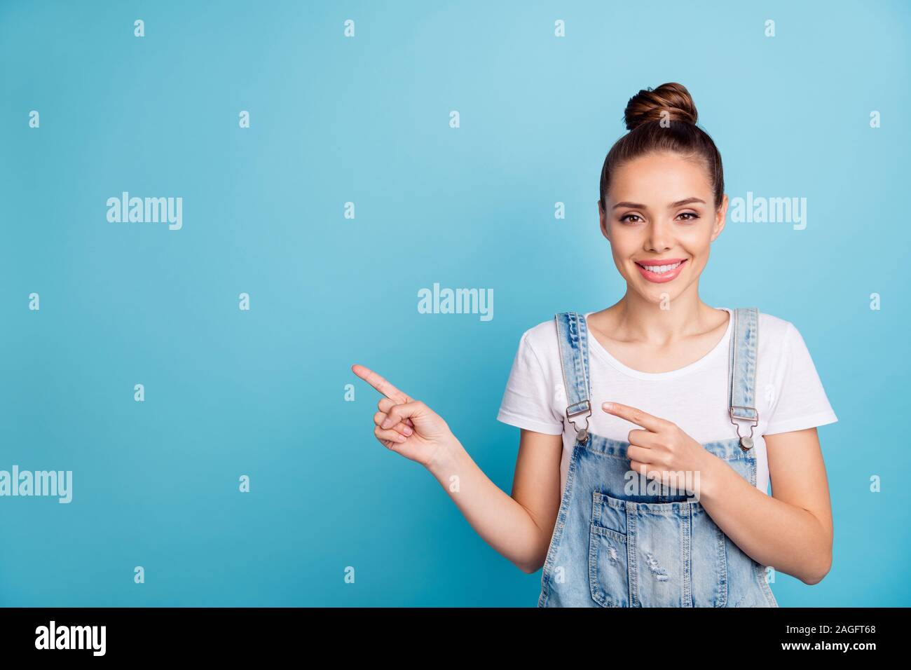 Portrait of lovely girl pointing at copy space wearing white t-shirt denim jeans overalls isolated over blue background Stock Photo
