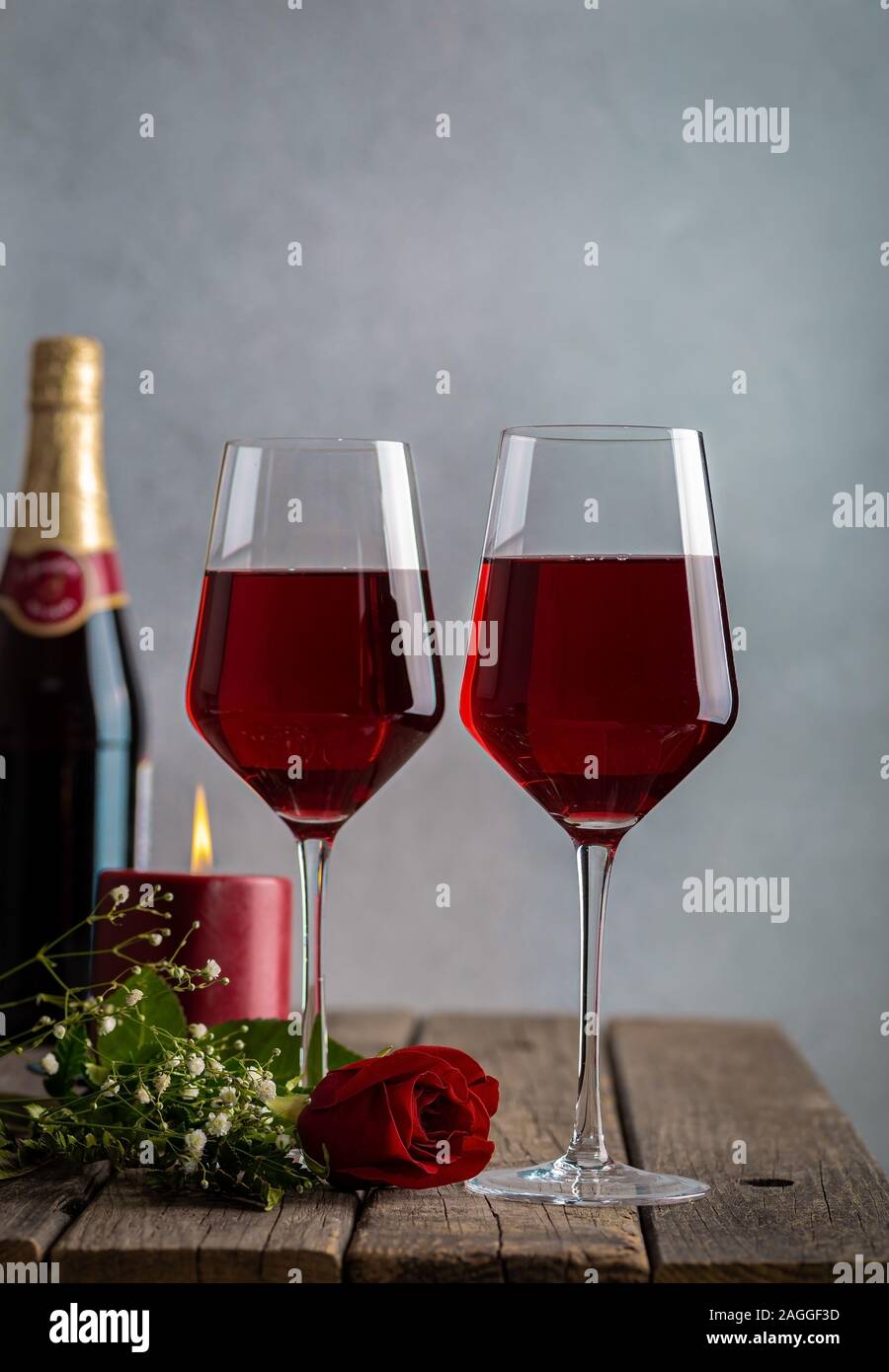 Two glasses of wine and red rose with burning candle and bottle in background on rustic wooden table Stock Photo