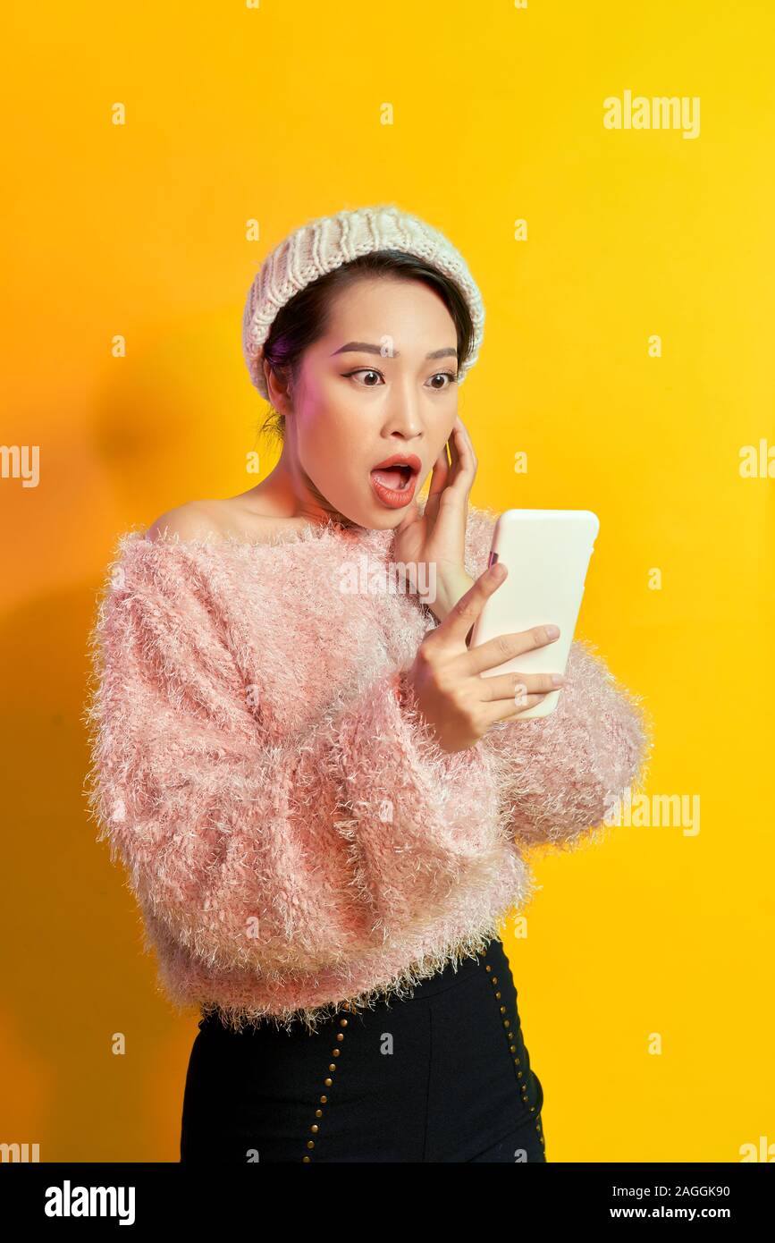 Shocked girl looking at phone screen with mouth open. Outdoor portrait of surprised young woman  wearing pink fur attire and holding smartphone. Stock Photo