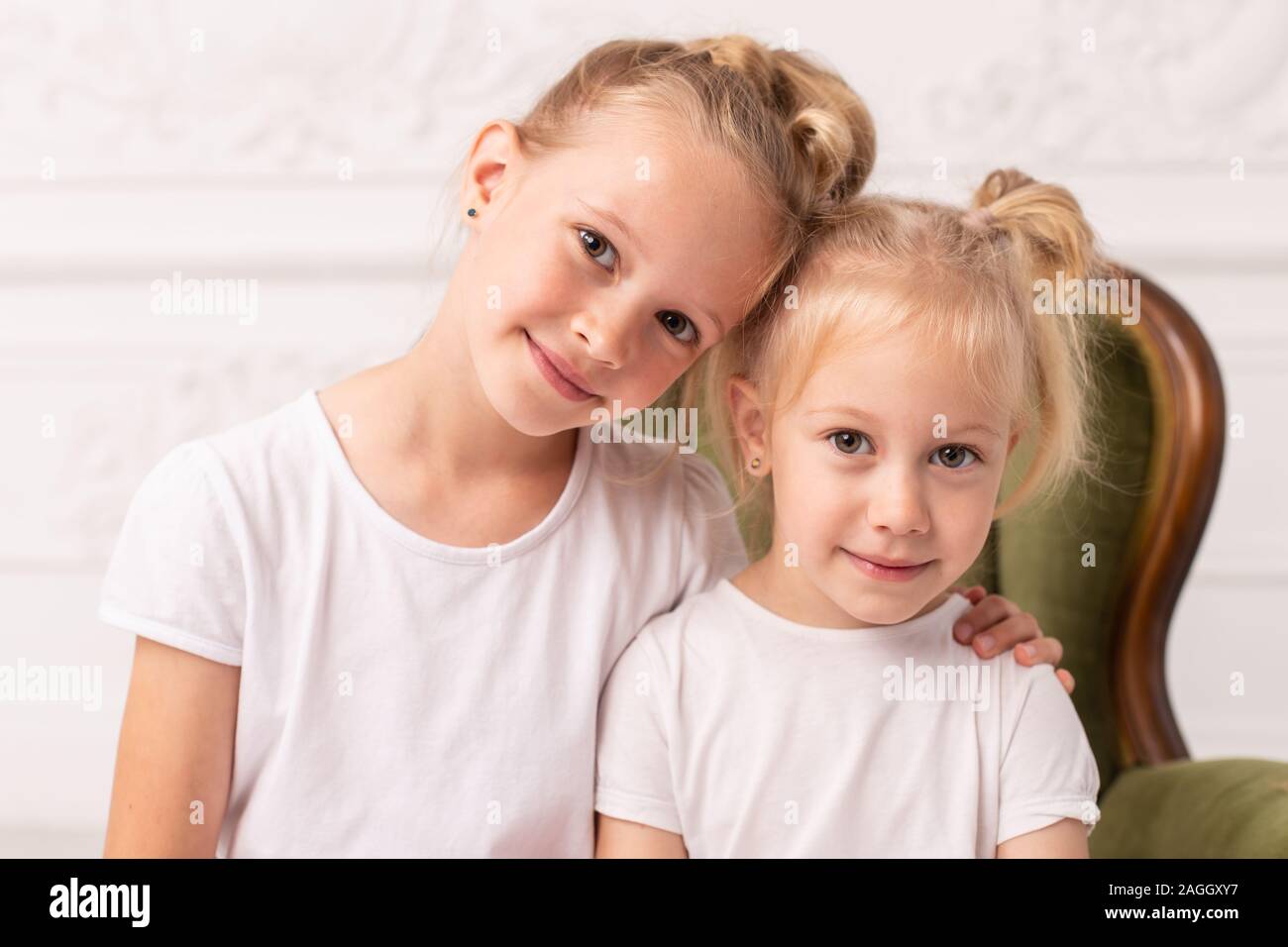 Two little blond smiling girls pose for camera while sit in green wooden armchair. Stock Photo
