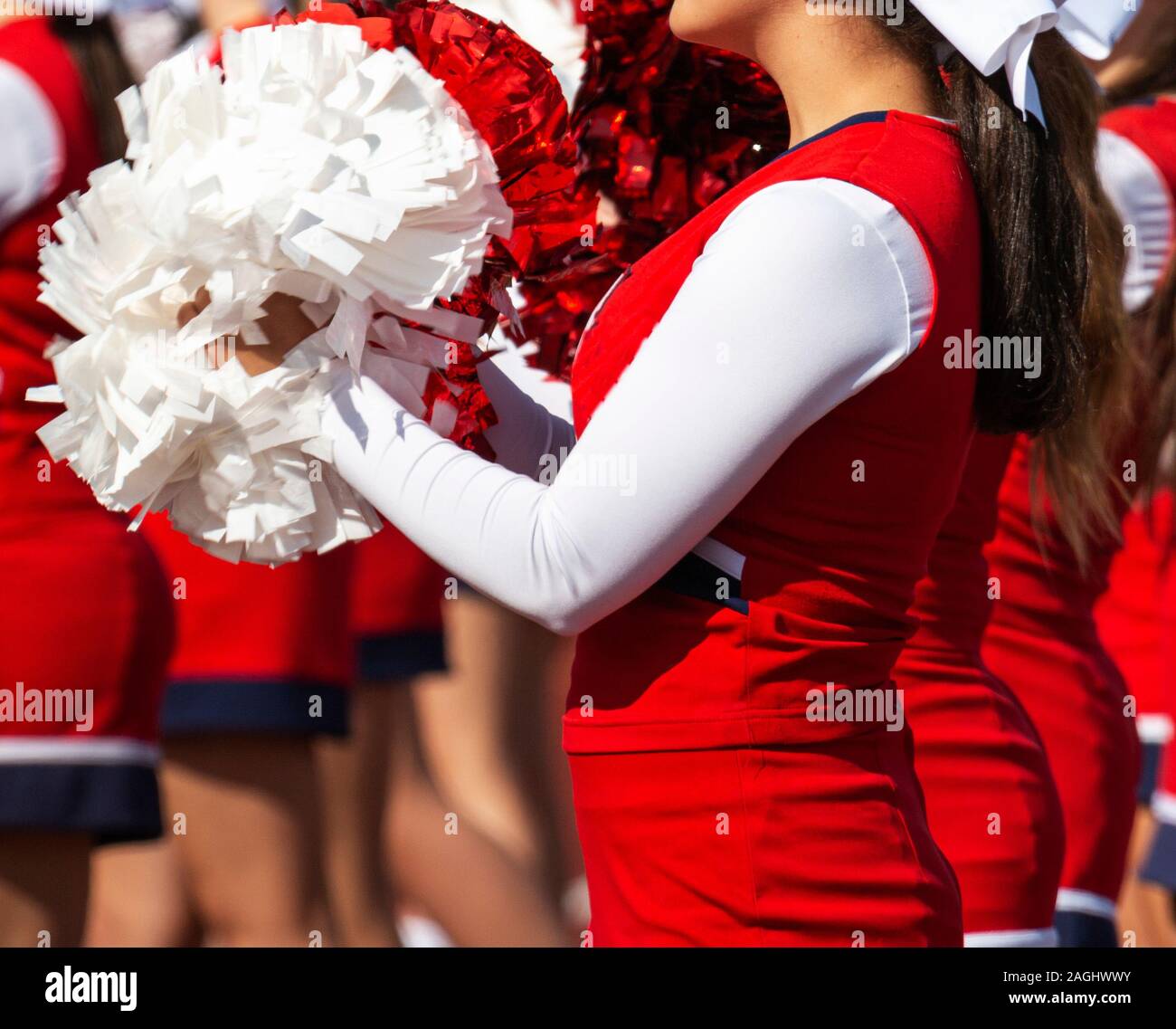 A high school cheerleader is facing the stands cheering for her team with red and white pom poms. Stock Photo