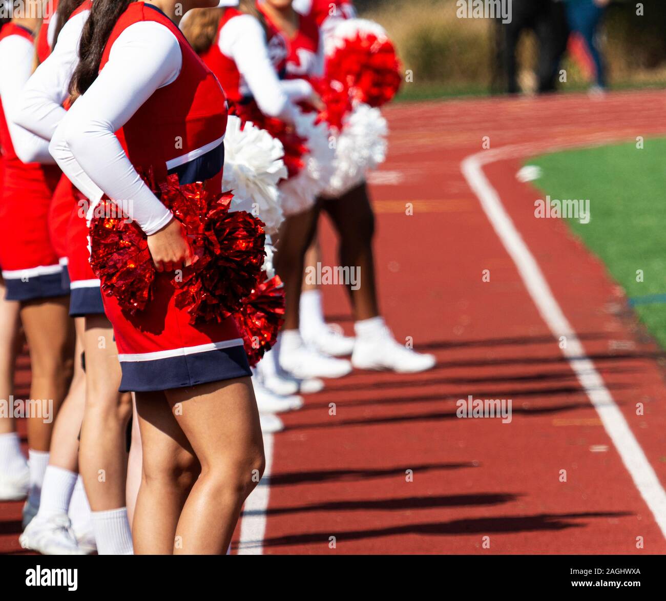 High school cheerleaders standing on the track holding red and white pom poms while watching the football game. Stock Photo