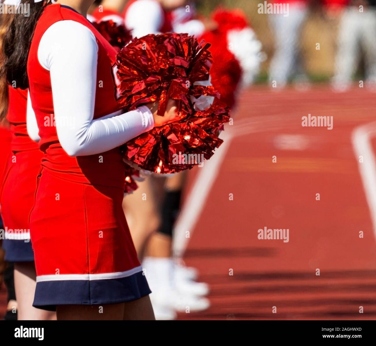 Sunlight reflects off of the red pom poms that cheerleaders are holding while standing on a track watching a high school football game. Stock Photo