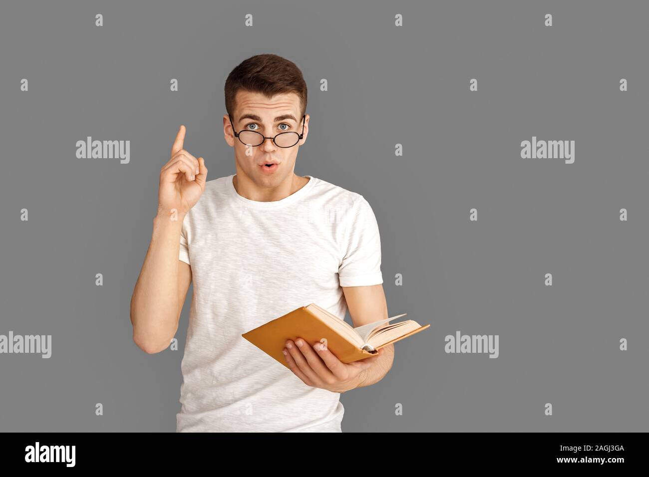 Freestyle. Young student in eyeglasses standing isolated on grey with book pointing up idea Stock Photo