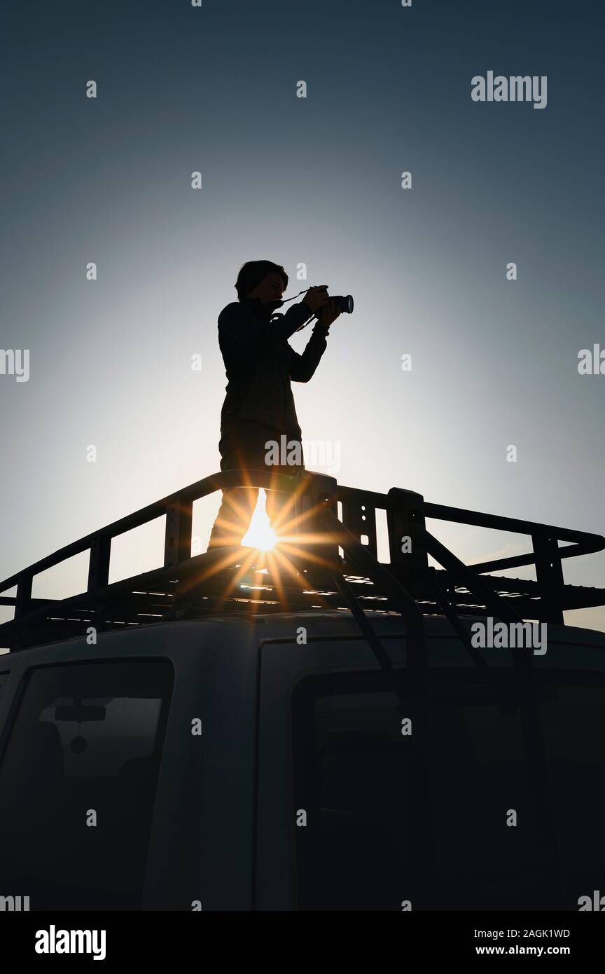 Female photographer takes pictures standing on car roof rack. Contre jour picture. Image for travel and adventure layouts Stock Photo