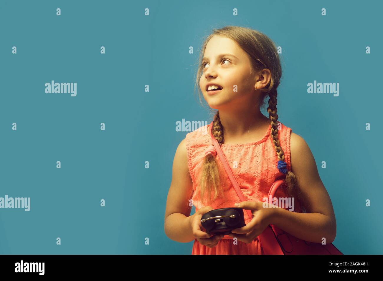 Back to school and childhood concept. Kid with distracted face expression.  Girl with black alarm clock and pink bag. Pupil with braids, isolated on  blue background with copy space Stock Photo -