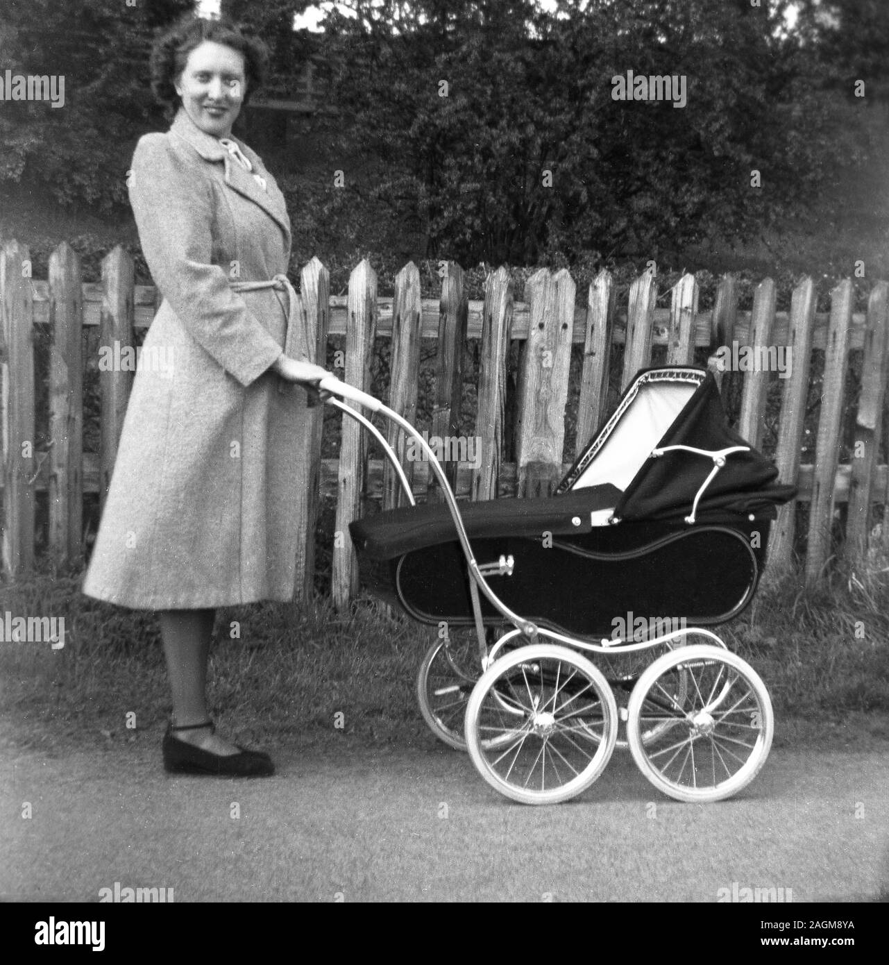 1950s, historical, a lady in a coat standing outside holding the handle of a traditional four-wheeled coach-built baby carriage or pram of the era, England, UK. Stock Photo