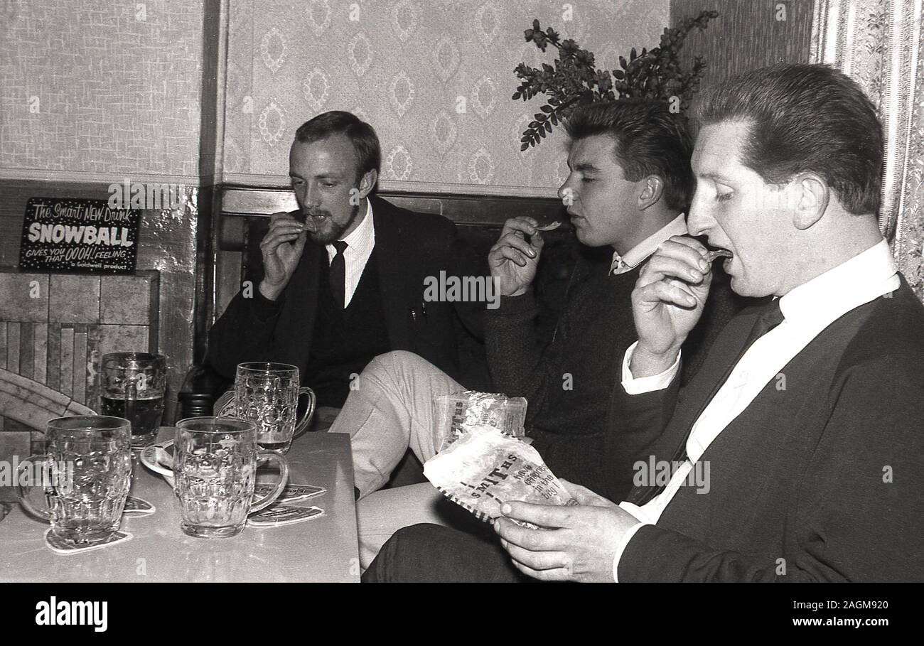 1970s, historical, three men sitting in a corner of a bar or pub eating crisps and enjoying a tankard of beer, England, UK. A sign for the latest drink, a 'snowball' can be seen on the fireplace mantle. A cocktail, a Snowball was  made with advocaat and lemonade. Stock Photo