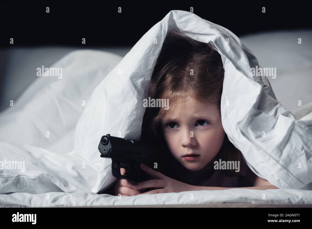frightened child holding gun while hiding under blanket isolated on