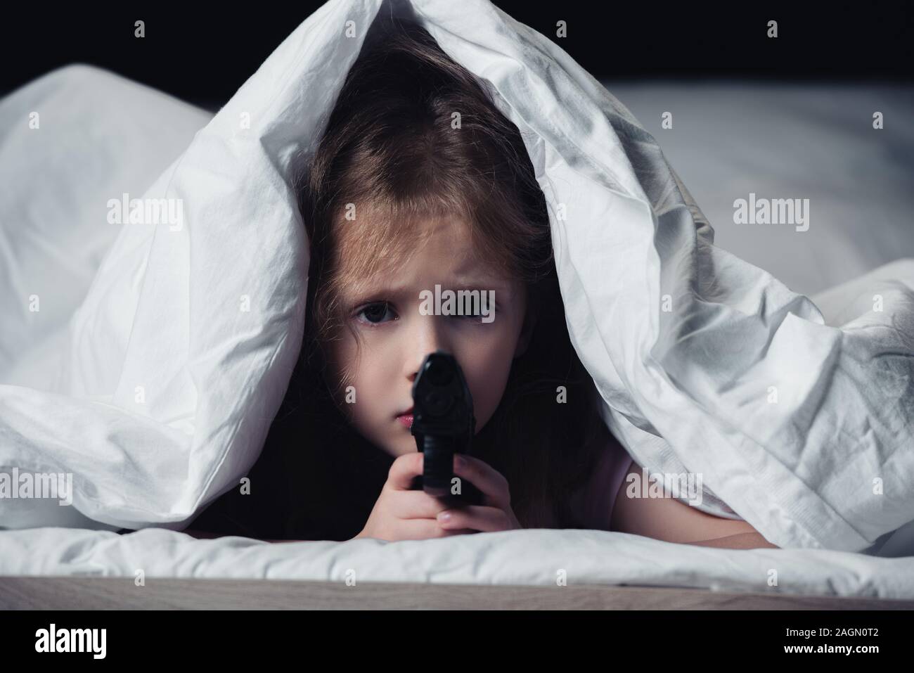 frightened child holding gun and looking at camera while hiding under blanket isolated on black