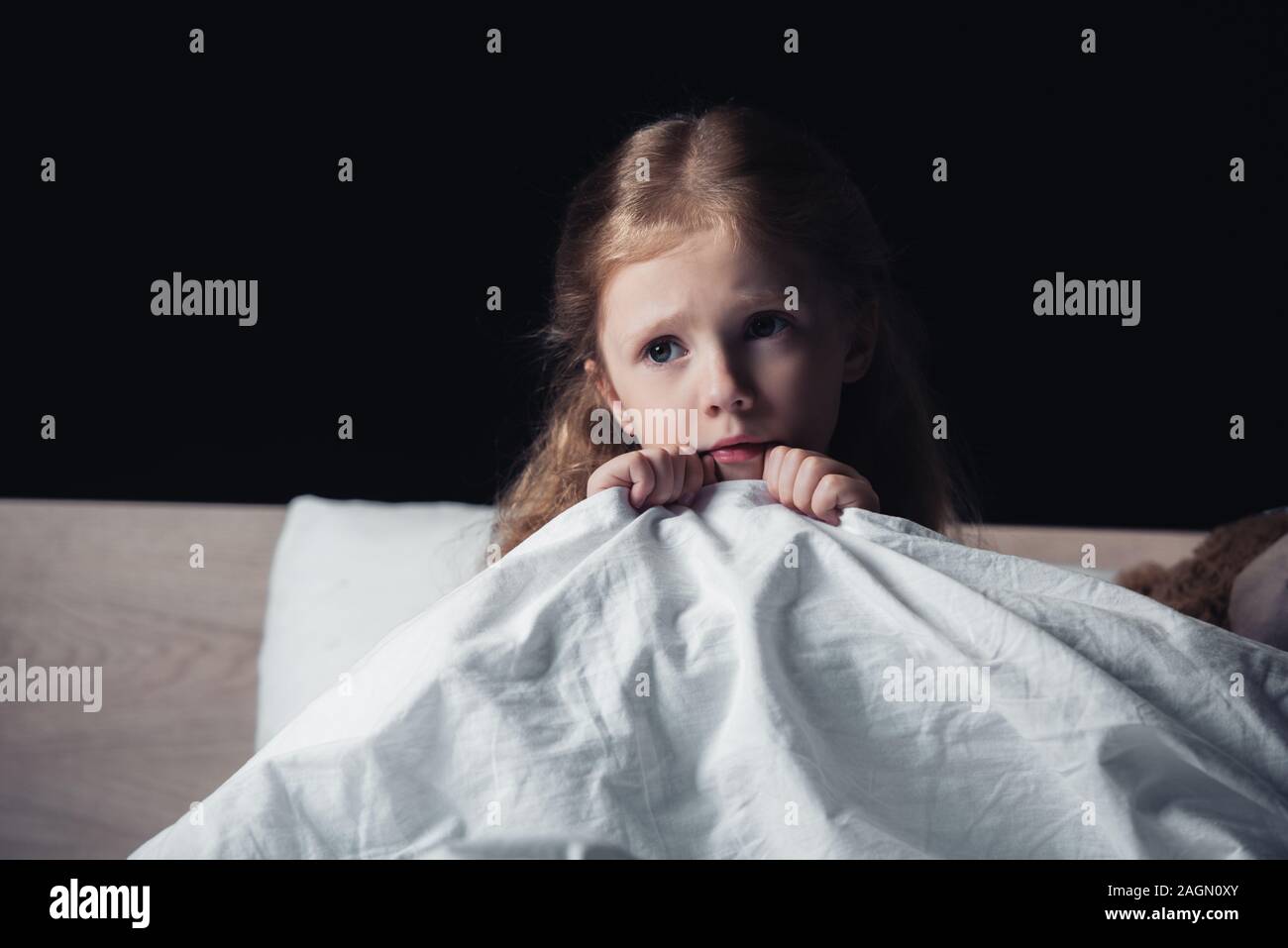 scared child looking away while sitting on bedding in dark bedroom isolated on black Stock Photo