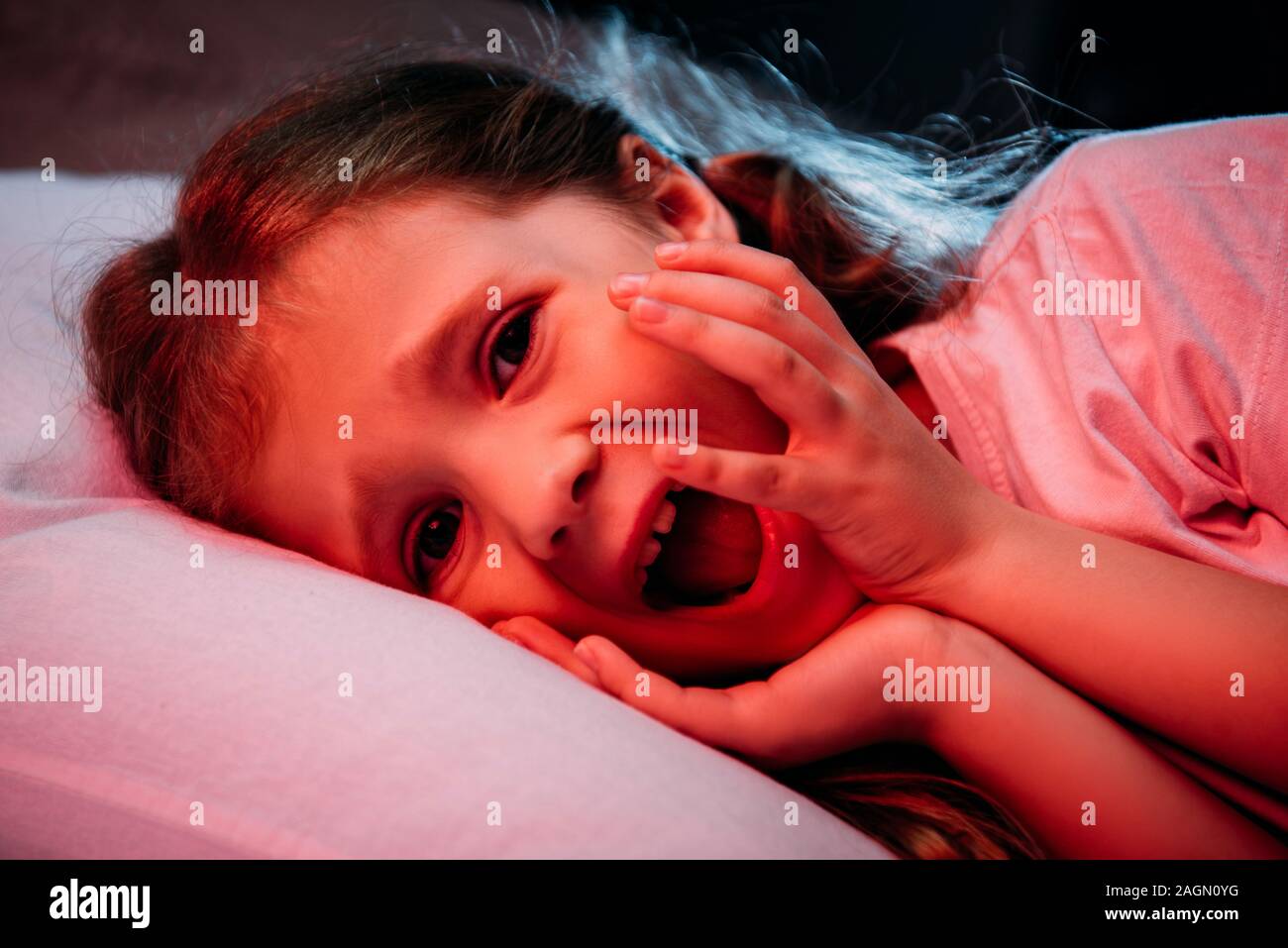frightened child screaming and looking at camera while lying in dark bedroom Stock Photo