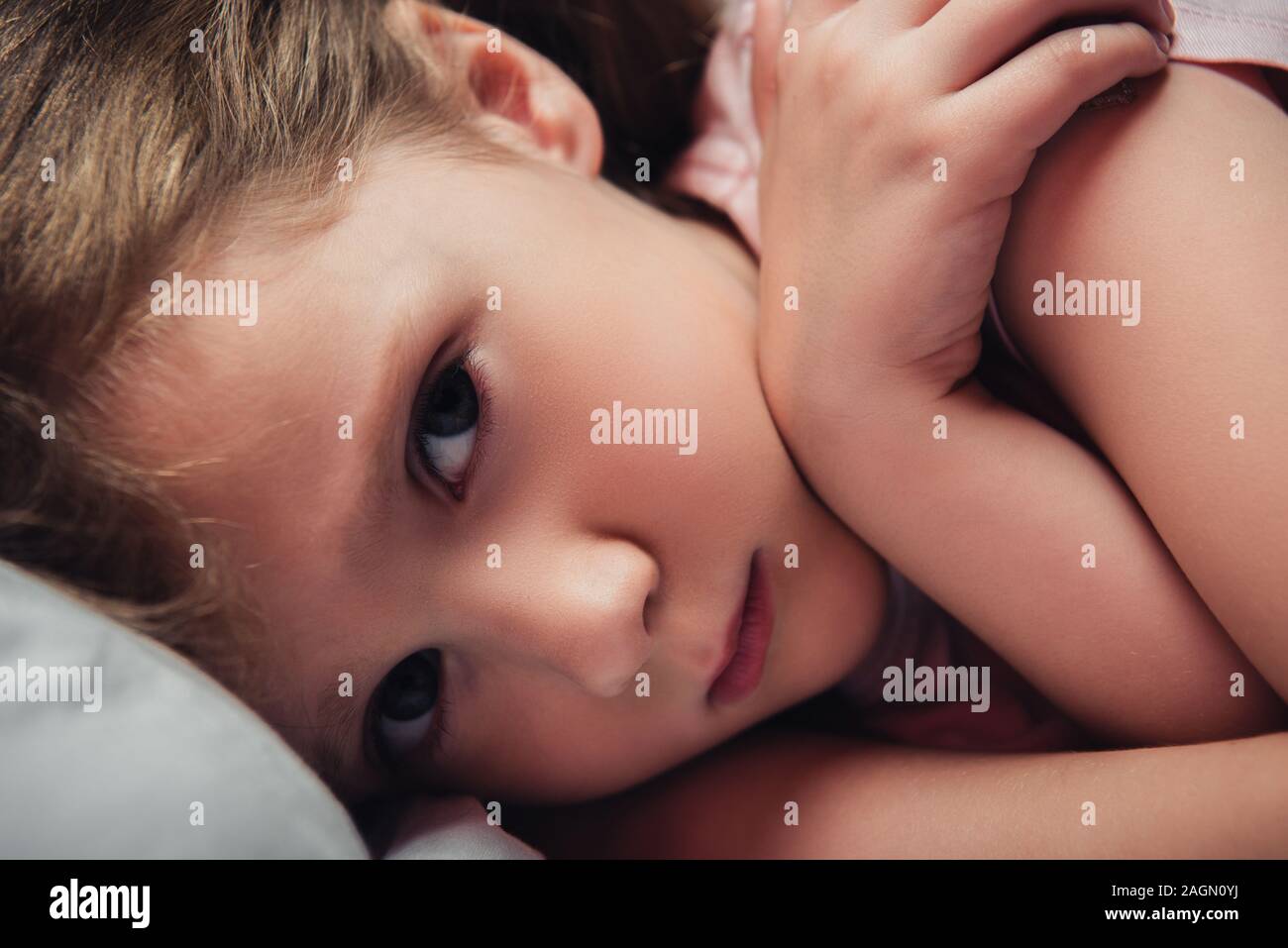 cute, frightened child looking at camera while lying in dark bedroom Stock Photo