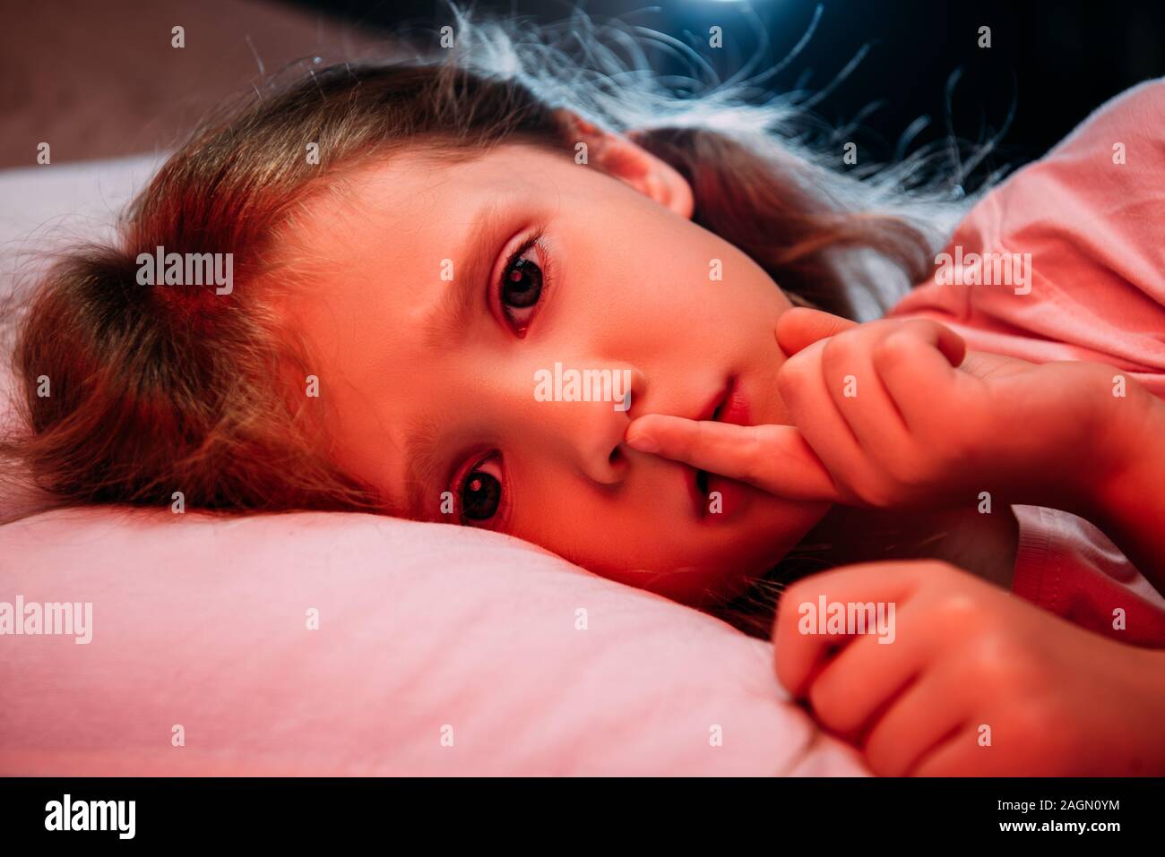 scared child lying in dark bedroom, showing hush sign and looking at camera Stock Photo