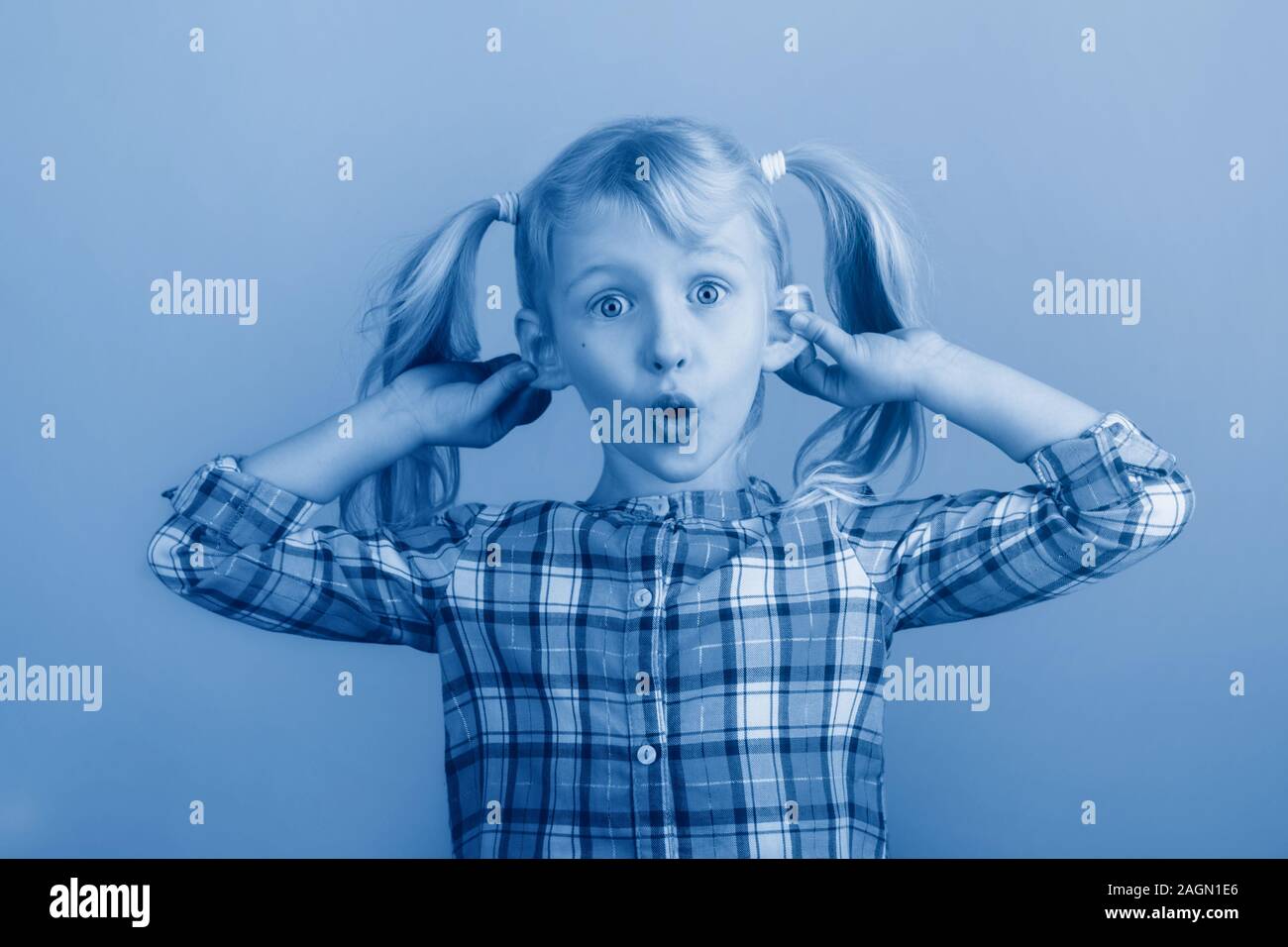 Funny Caucasian young girl making faces in front of camera. Child holding pulling dragging ears. Kid expressing emotions. April fool day concept. Tone Stock Photo