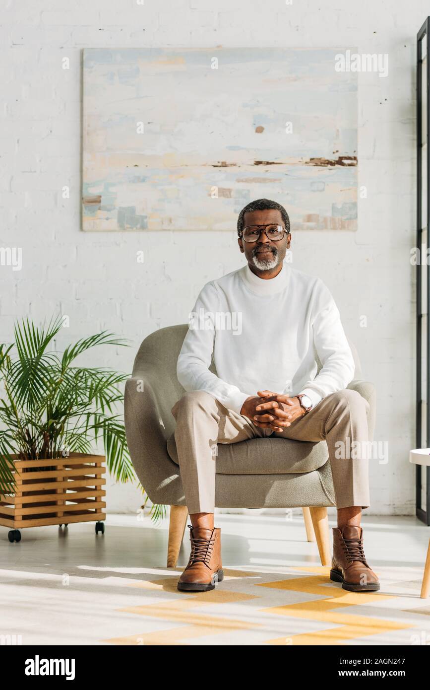 stylish african american man sitting in armchair and looking at camera ...