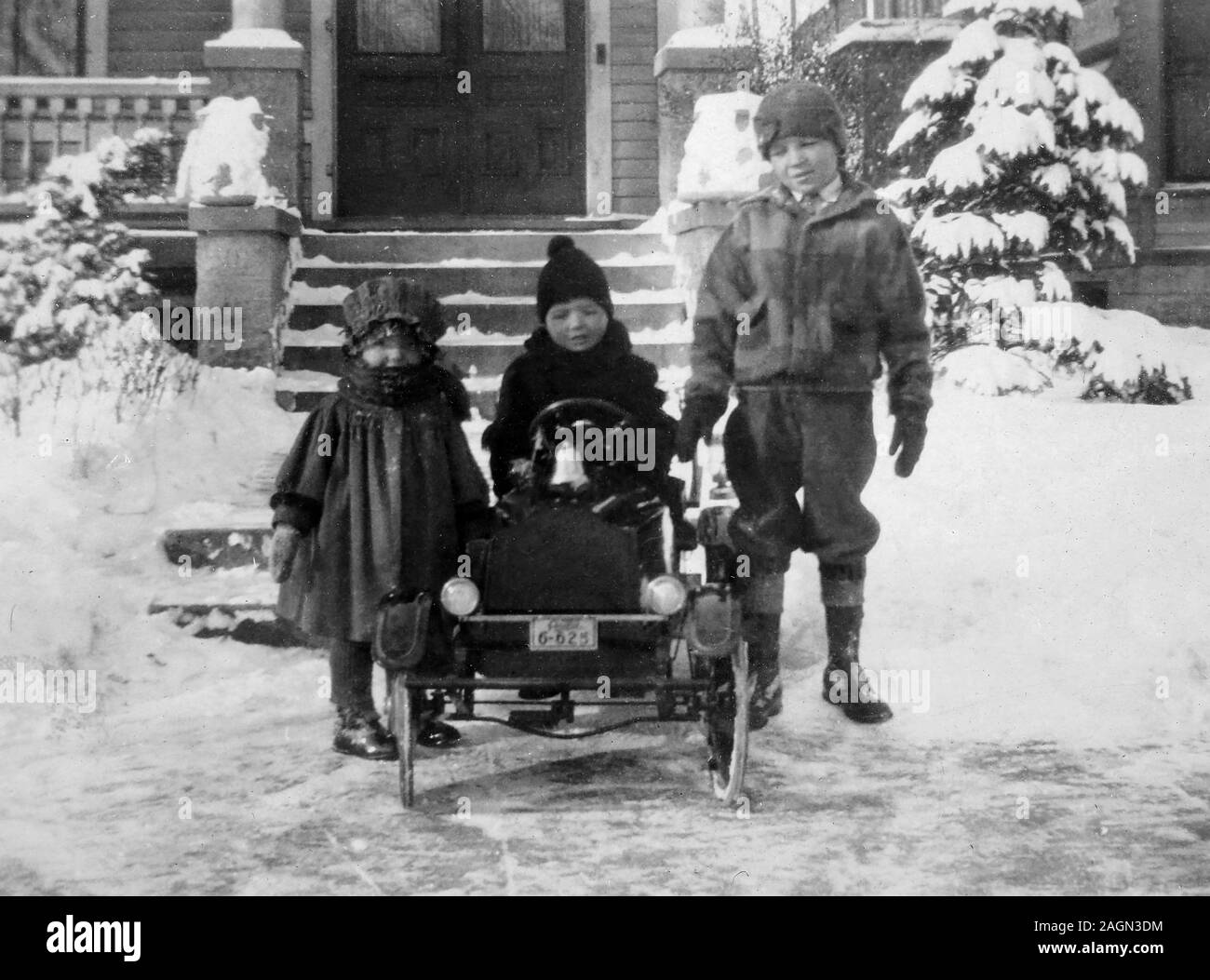 Three children bundled up for a cold day play with their period toy automobile, ca. 1925. Stock Photo