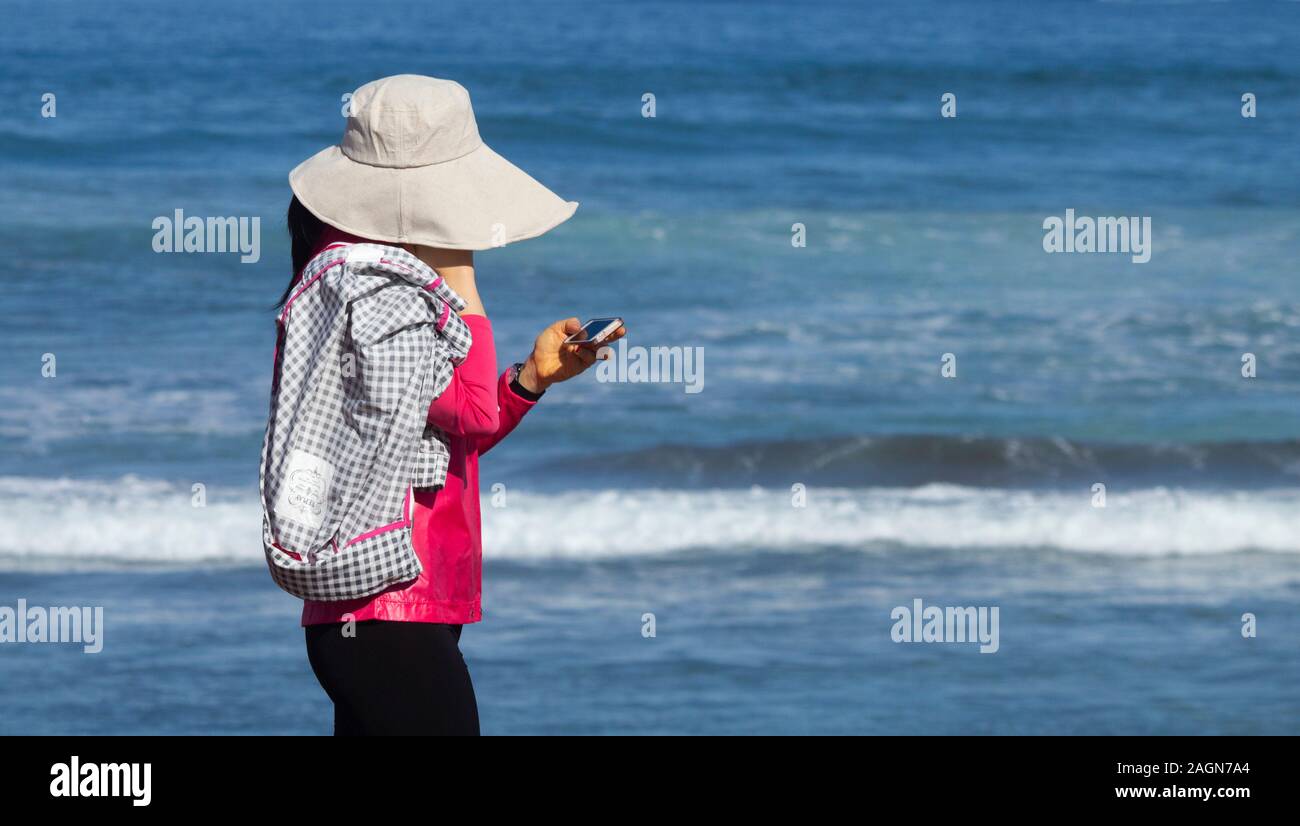 Woman wearing hat for sun protection looking at mobile phone while walking on beach. Stock Photo