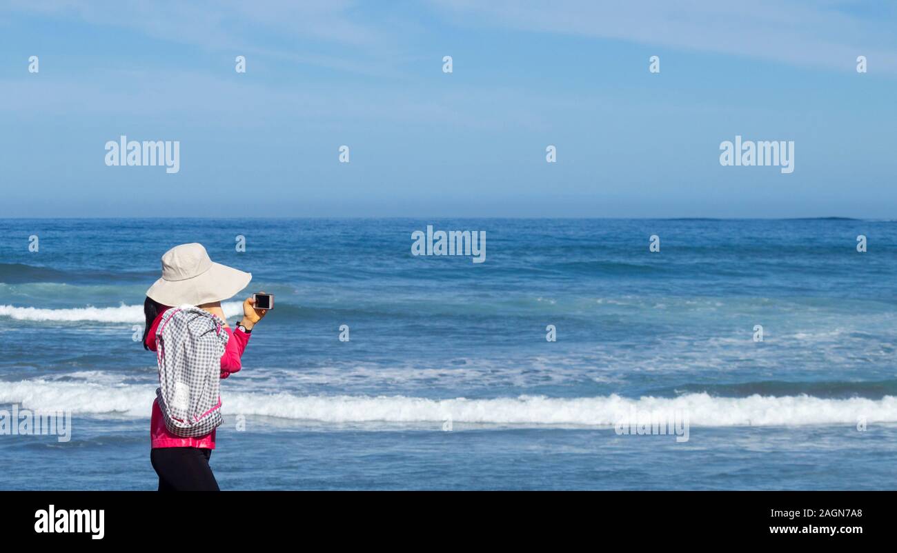 Woman wearing hat for sun protection looking at mobile phone while walking on beach. Stock Photo