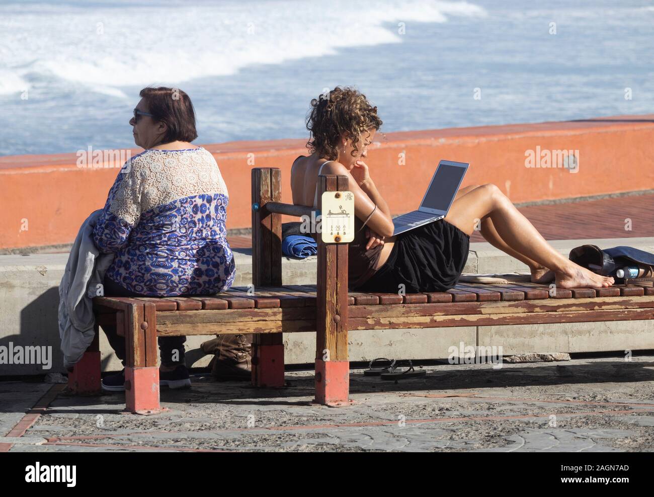 Younger woman on beach sitting on gym bench looking at laptop while elderly woman looks out to sea. Stock Photo