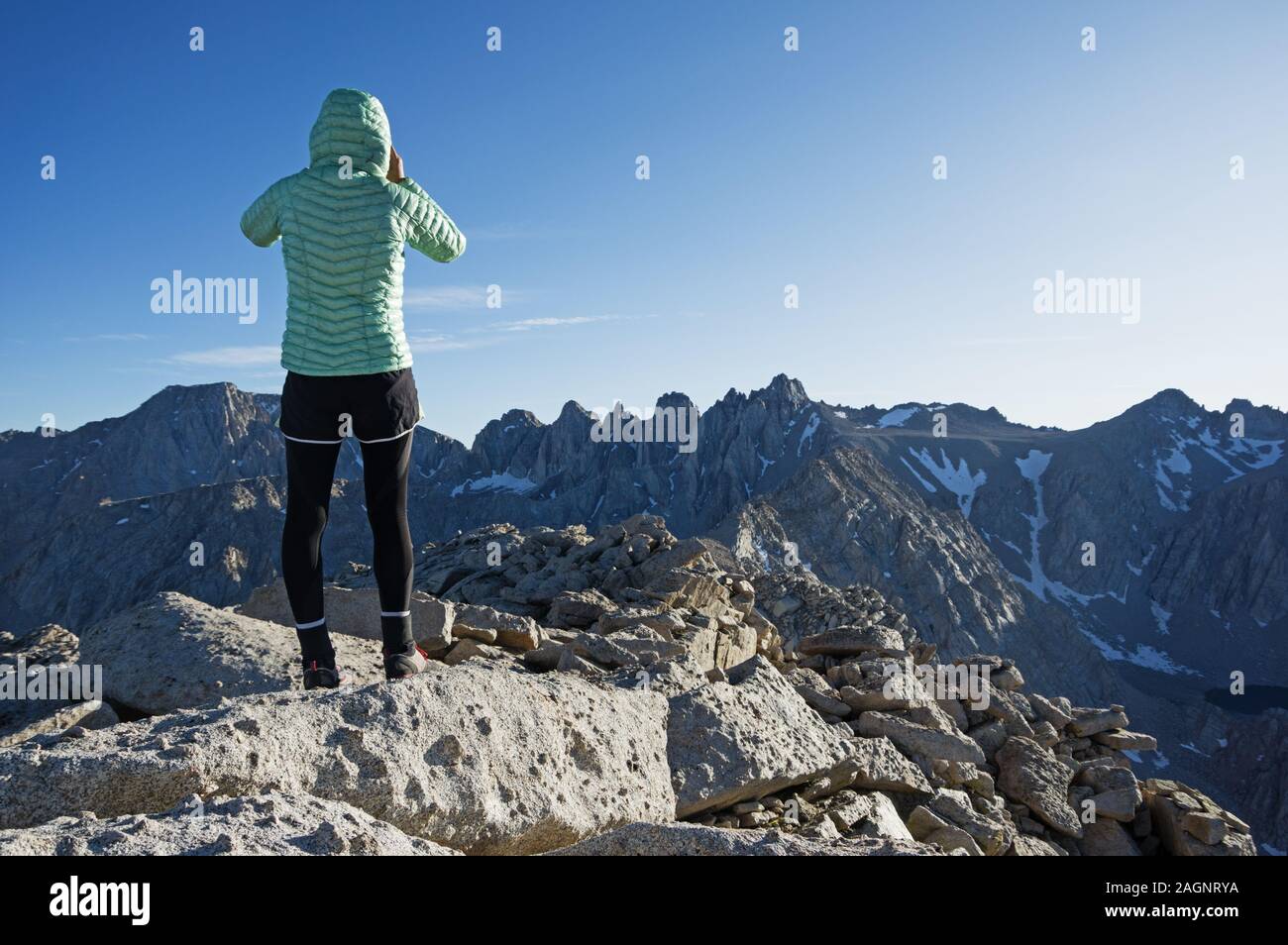 back view of a woman in a puffy jacket and tights taking a photo from Lone Pine Peak towards Mount Langley and Comb Ridge Stock Photo