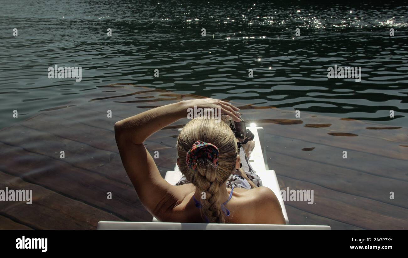 Woman shooting on handheld film gimbal stabilization for smartphone. Girl lie sunbed on pier make selfie. Blogger broadcast video vlogging. Stock Photo