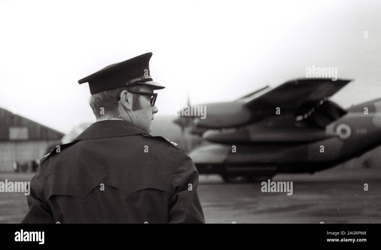 A black and white image, taken in the 1970's, of a male security guard at an airfield in the uk with an out of focus propellor plane in the background Stock Photo