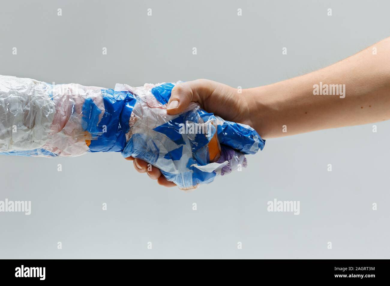 Big plastic hand made of garbage shaking another hand on white studio background. Polymers overusing and overproduction. Ecology problems, pollution, recycling. It's getting dangerous for humanity. Stock Photo